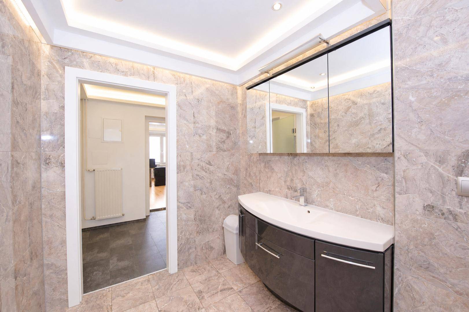Modernes Badezimmer mit Waschtisch, Spiegelschrank und Zugang zu einem angrenzenden Raum.