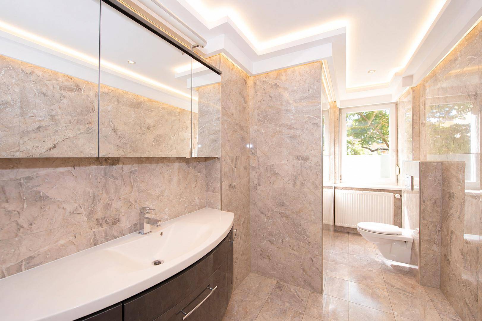 Modernes Badezimmer mit Marmorfliesen, Waschtisch, Spiegelschrank und indirekter Beleuchtung.