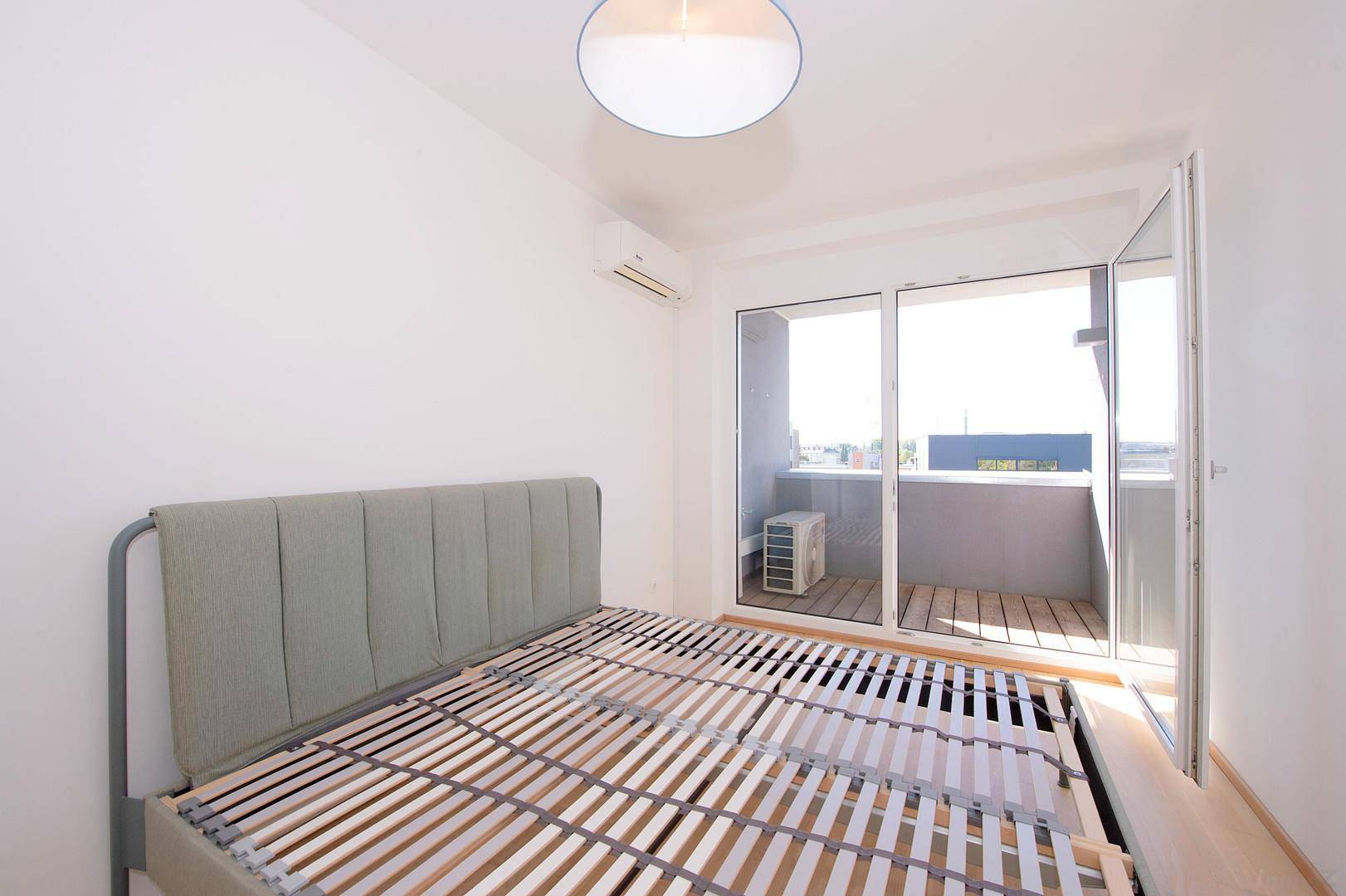 Schlafzimmer mit Bettrahmen, Klimaanlage und großer Balkontür für viel Tageslicht.