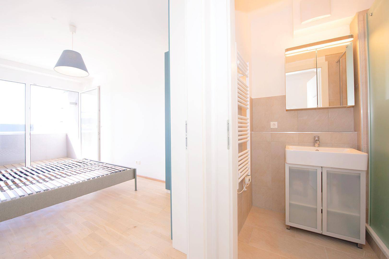 Blick vom Badezimmer in ein Schlafzimmer mit Bettrahmen und Balkonzugang.