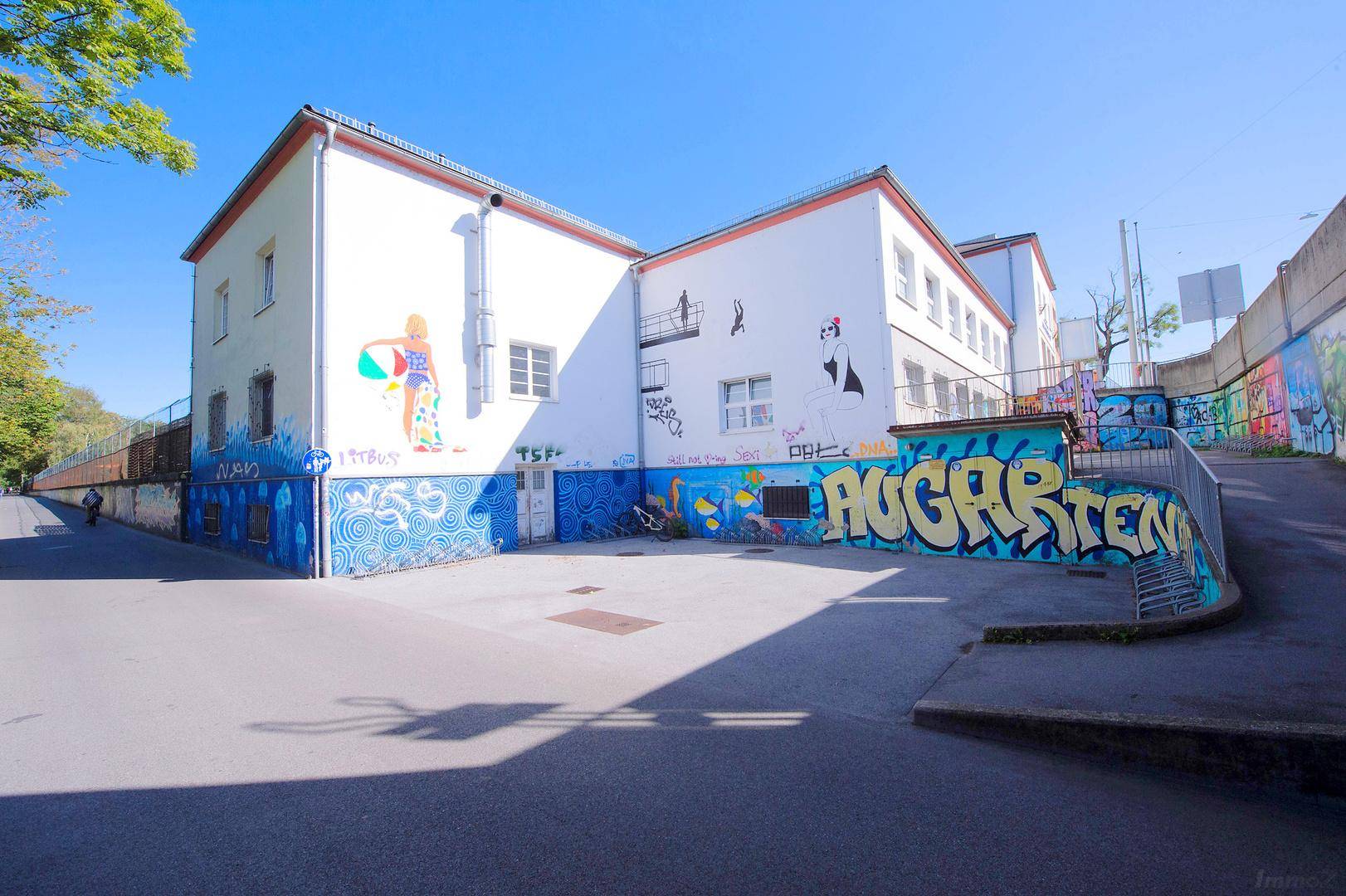 Gebäudeaußenansicht mit farbenfrohen Wandmalereien und Graffiti, umgeben von einem sonnigen Platz.