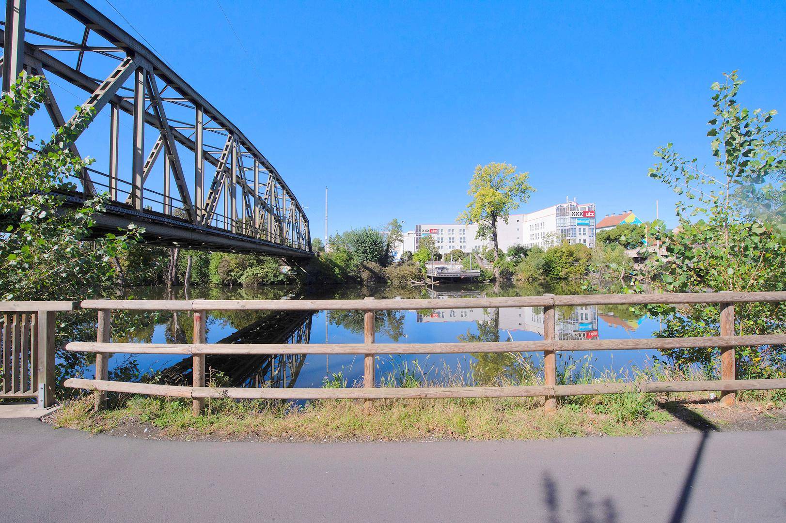 Ruhiger Flusslauf mit Holzzaun im Vordergrund, einer Brücke und Gebäuden am Ufer.