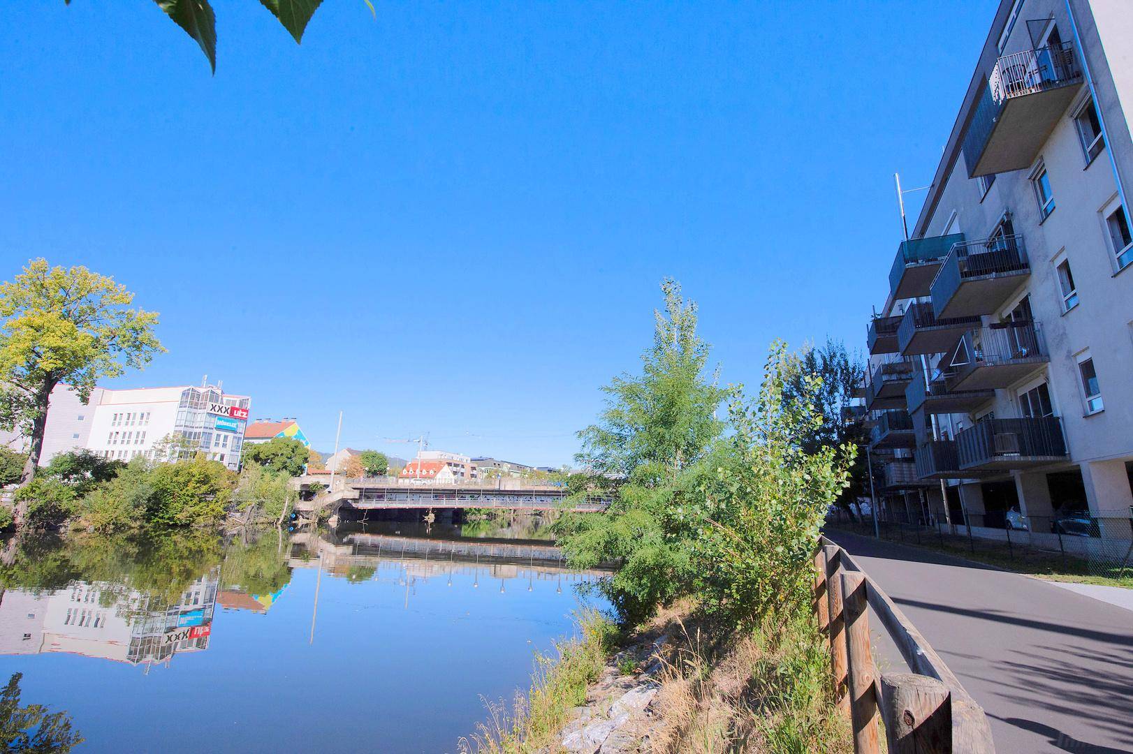 Uferpromenade entlang eines Flusses mit Bäumen und einem Wohngebäude mit Balkonen in der Ferne.