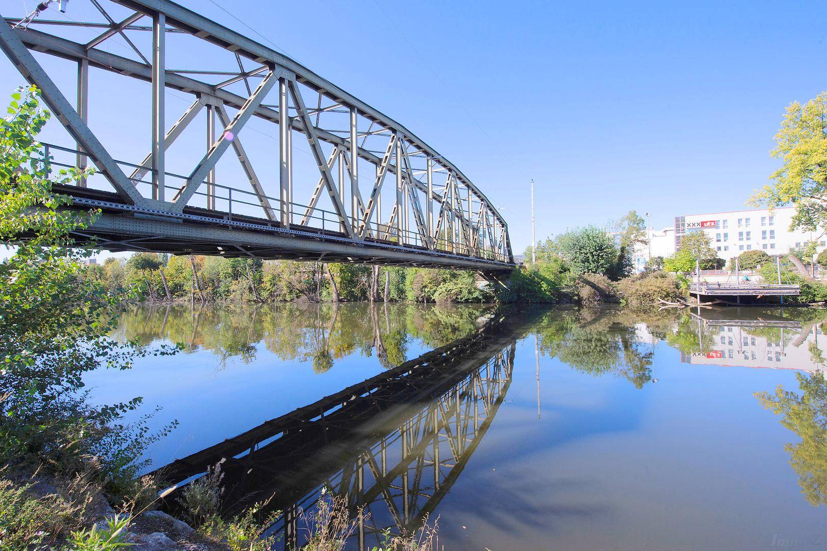 Flusslandschaft mit einer markanten Metallbrücke und Gebäuden im Hintergrund unter blauem Himmel.