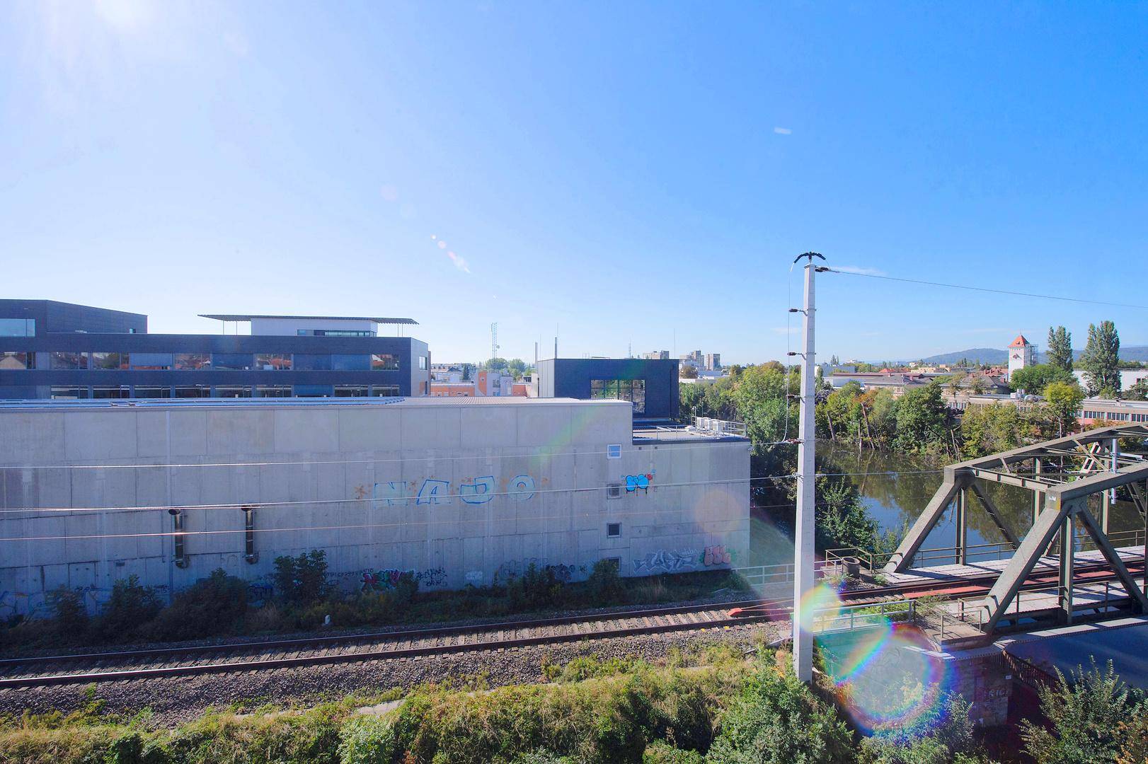 Weitblick über Fluss, Bahngleise und umliegende Gebäude unter blauem Himmel.