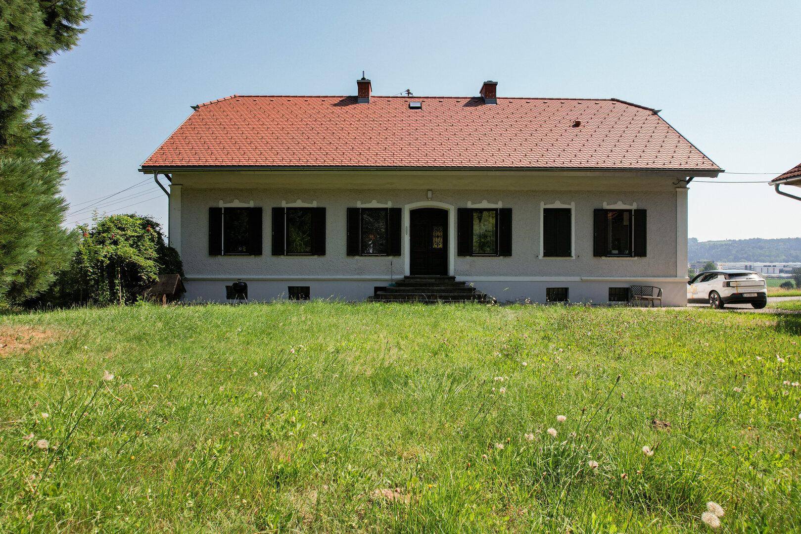 Vorderansicht eines traditionellen Hauses mit rotem Ziegeldach und grünen Fensterläden, umgeben von Rasen.