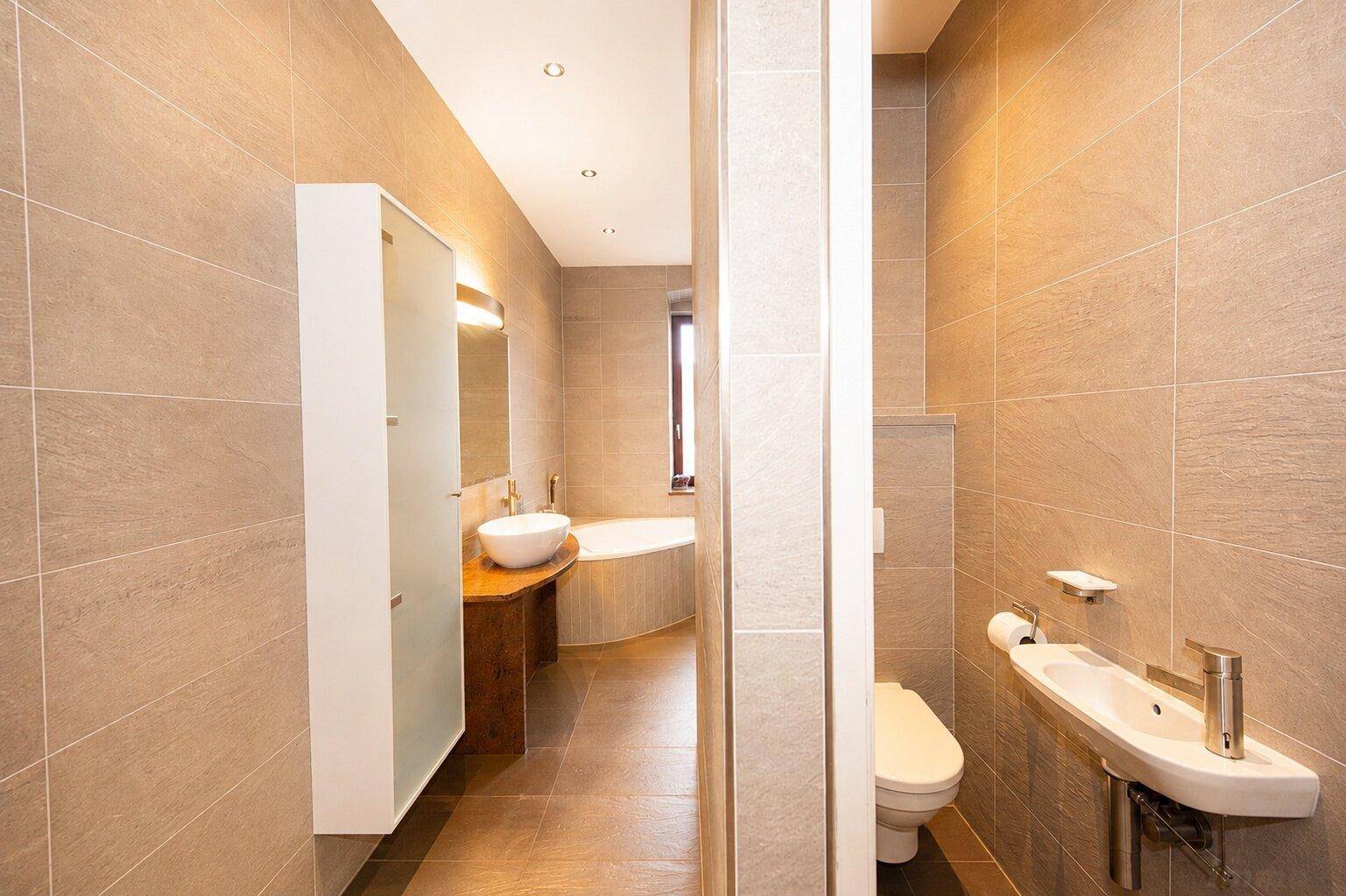 Modernes Badezimmer mit grauen Fliesen, separatem WC-Bereich und Blick auf eine Badewanne.