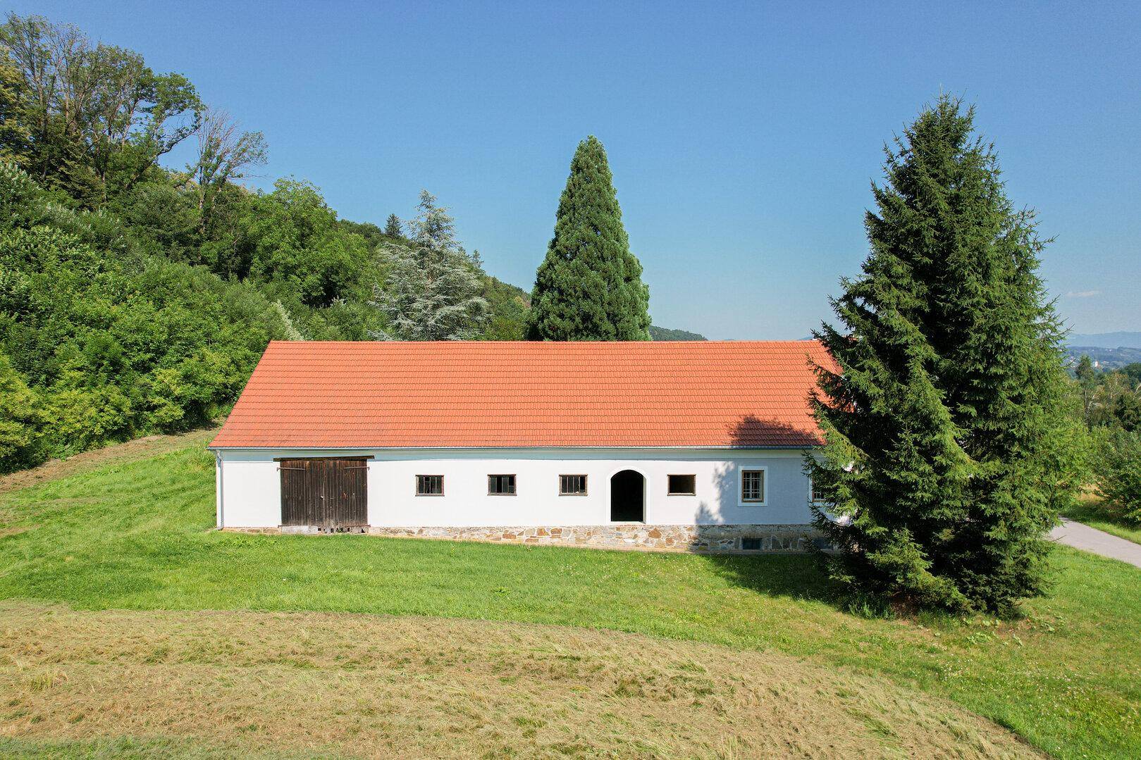 Nebengebäude mit Ziegeldach und Holztüren, umgeben von Bäumen und Wiesen.