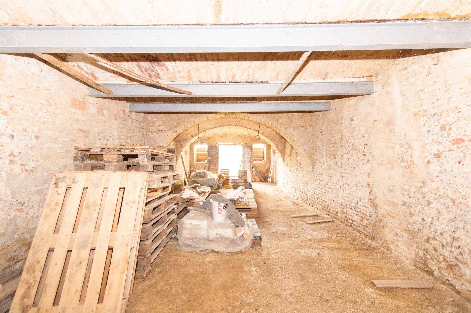 Unausgebauter Kellerraum mit Ziegelwänden, Holzbalken und gelagerten Paletten.