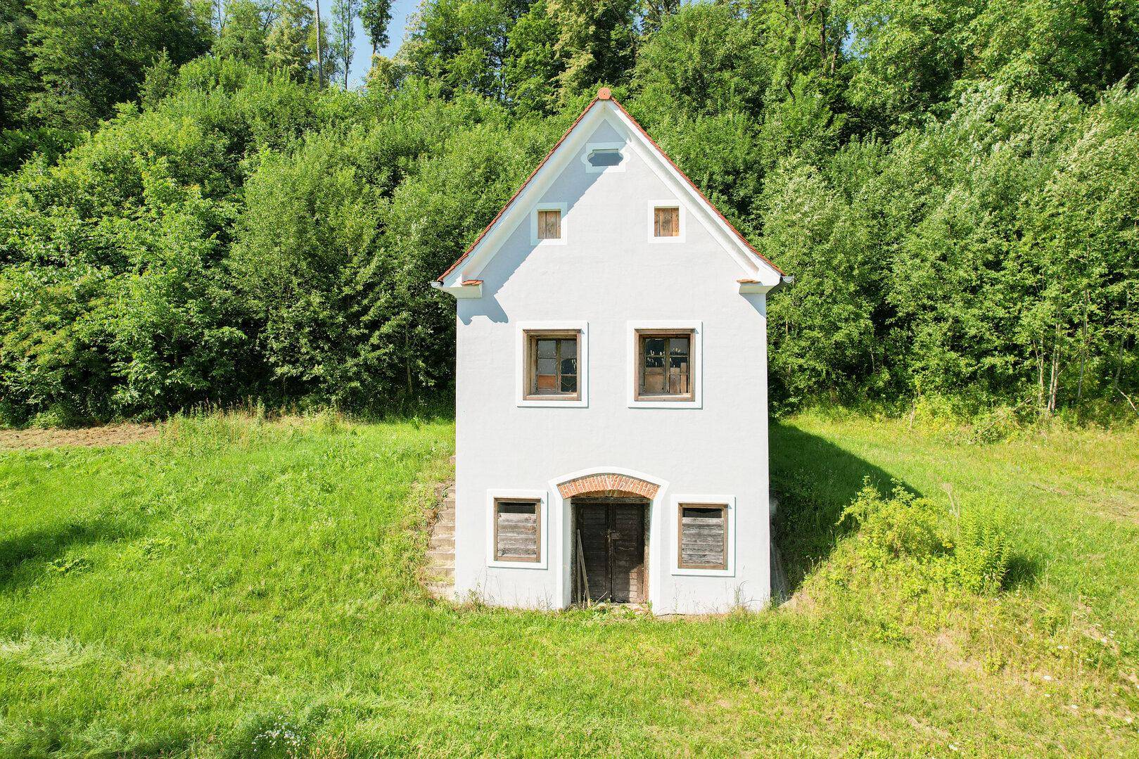 Kleines weißes Haus mit rotem Satteldach und Holztüren, umgeben von grüner Natur.