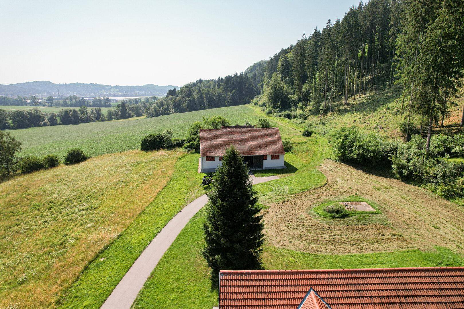 Weitläufige Landschaft mit einem Gebäude in einem Tal, umgeben von Wald und Feldern.