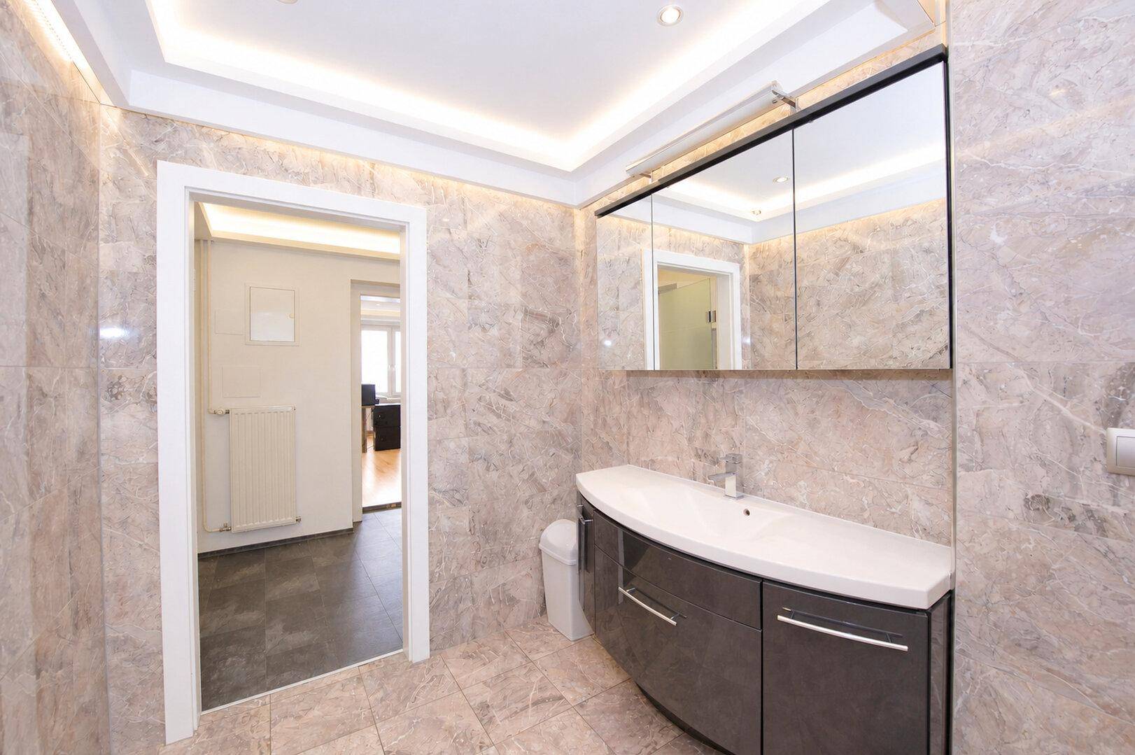 Modernes Badezimmer mit Waschtisch, Spiegelschrank und hellen Marmorfliesen, Zugang zu einem weiteren Raum.