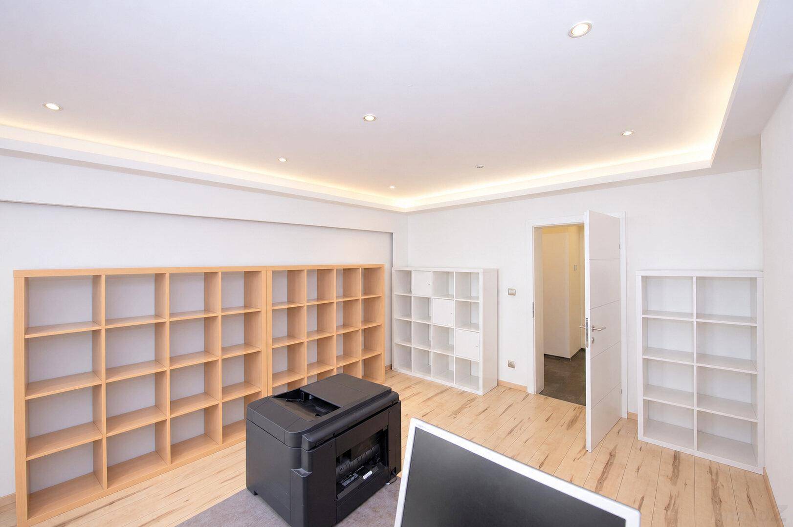 Helles Büro mit mehreren Regalen, Holzboden und einem Drucker auf dem Teppich.