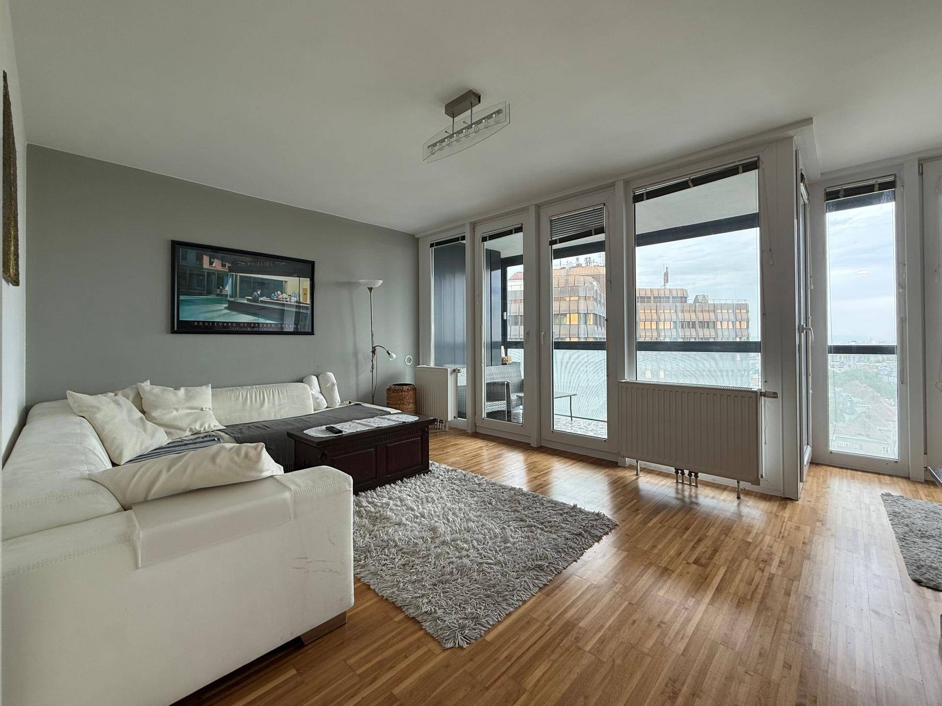 Geräumiges Wohnzimmer mit bequemer weißer Couch, Holzboden und weitem Blick über die Stadt.
