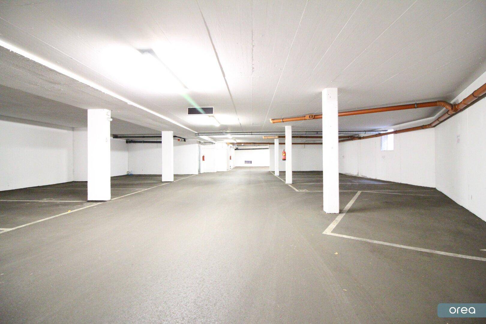 Leere und helle Tiefgarage mit klaren Stellplatzmarkierungen und sichtbaren Installationen an der Decke.