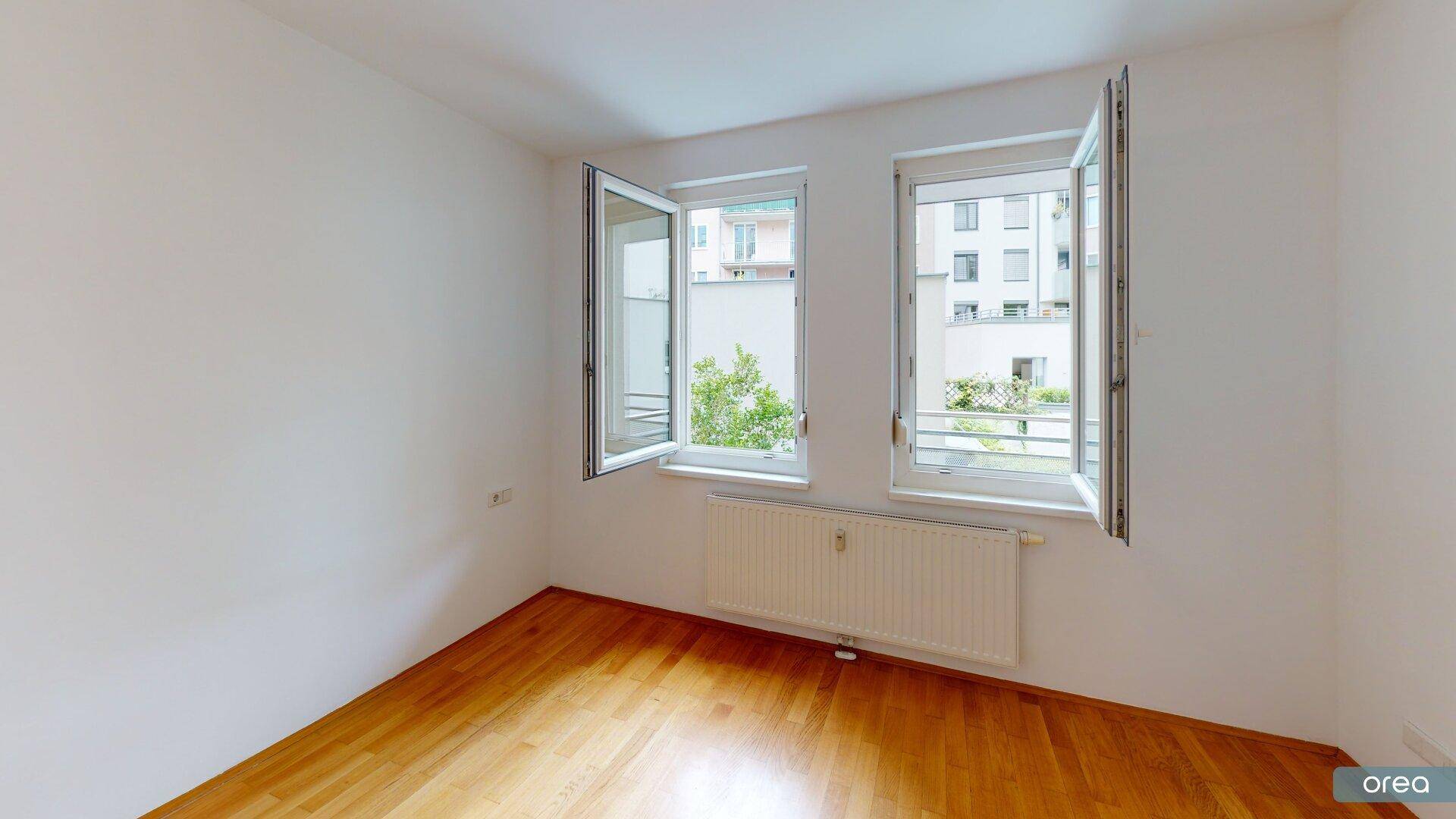 Helles Schlafzimmer mit Parkettboden und zwei großen Fenstern, die viel Tageslicht hereinlassen.