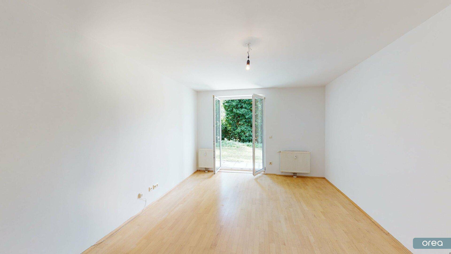 Heller, leerer Raum mit Parkettboden und direktem Zugang zum grünen Außenbereich.