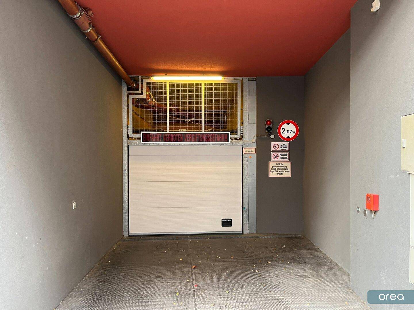Garageneinfahrt mit Rolltor, Höhenbegrenzungsschild und grauen Wänden unter einer roten Decke.