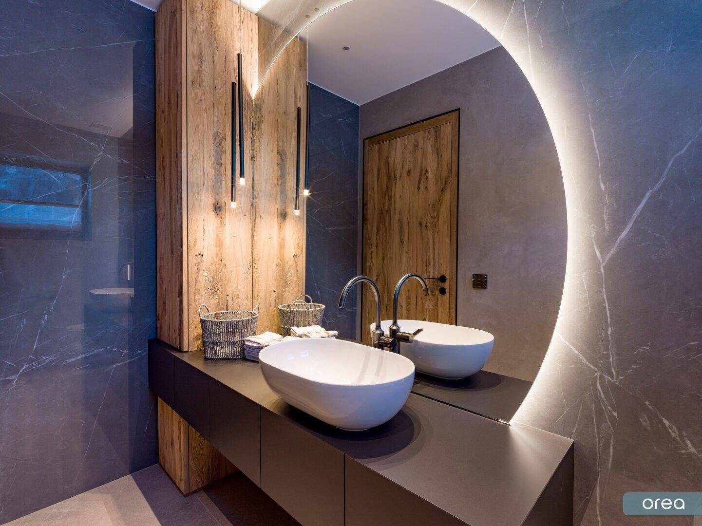 Elegantes Badezimmer mit runden beleuchteten Spiegeln, Doppelwaschbecken und dunklen Marmorfliesen.