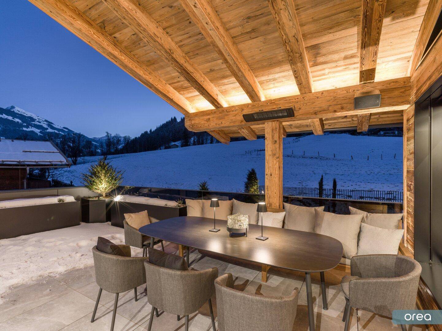 Überdachte Terrasse mit Esstisch und Sitzgelegenheiten, umgeben von verschneiter Landschaft.