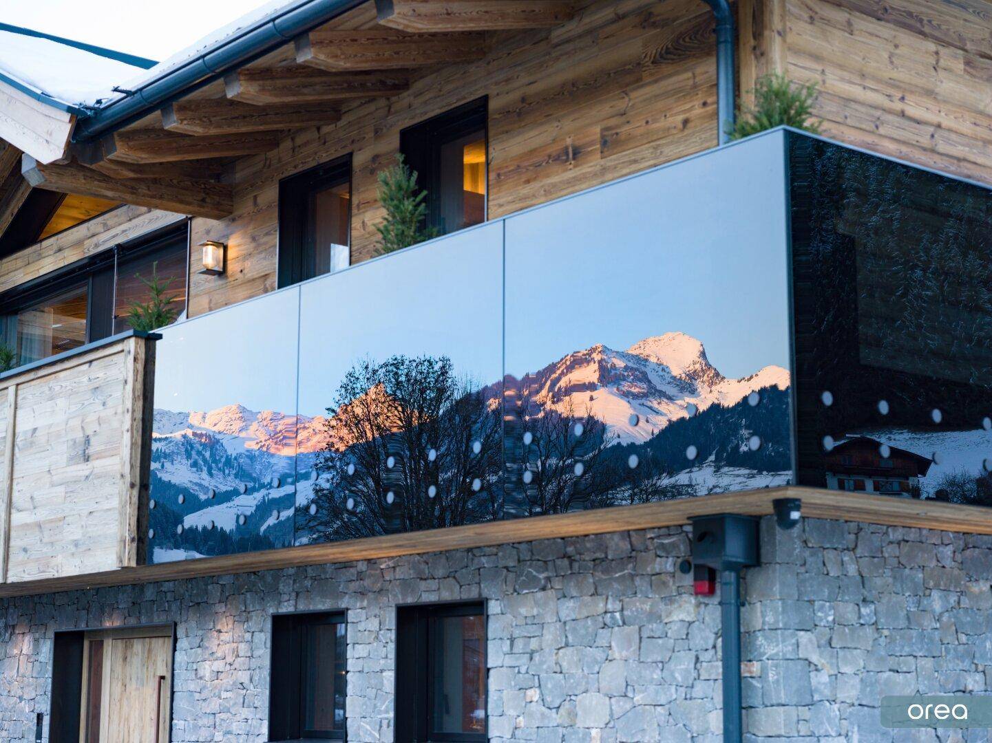 Detailansicht eines Balkons mit Glasbrüstung, die die schneebedeckten Berge widerspiegelt.