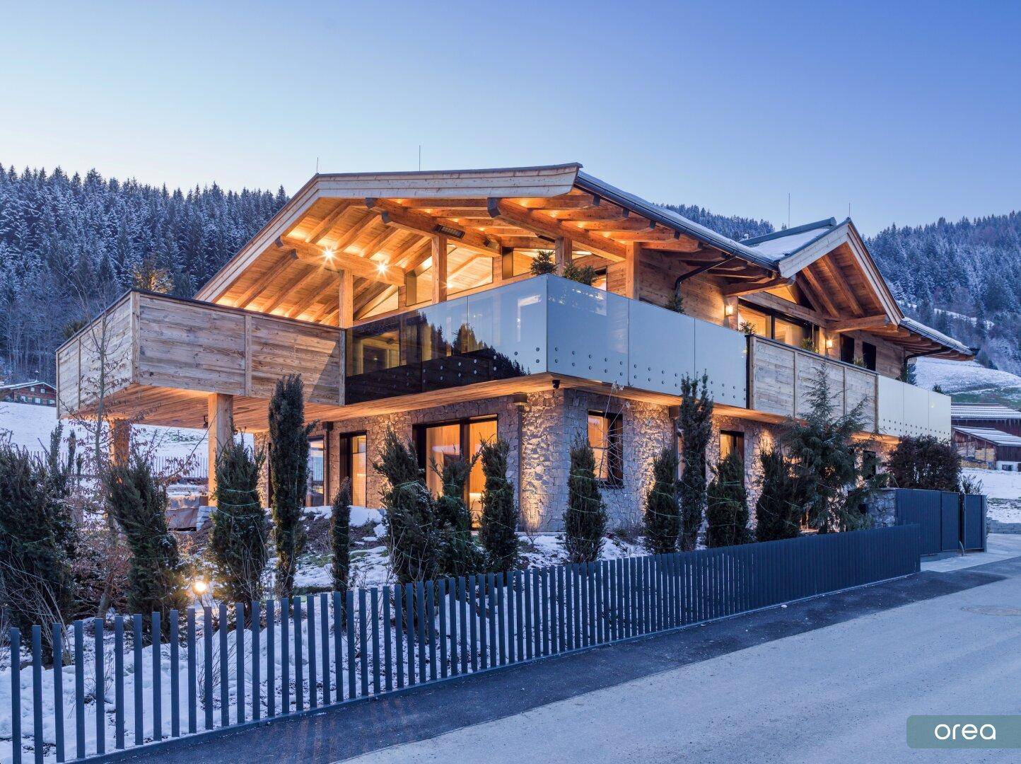 Modernes Chalet mit Holz- und Steinfassade, großen Glasflächen und beleuchteten Balkonen in verschneiter Landschaft.
