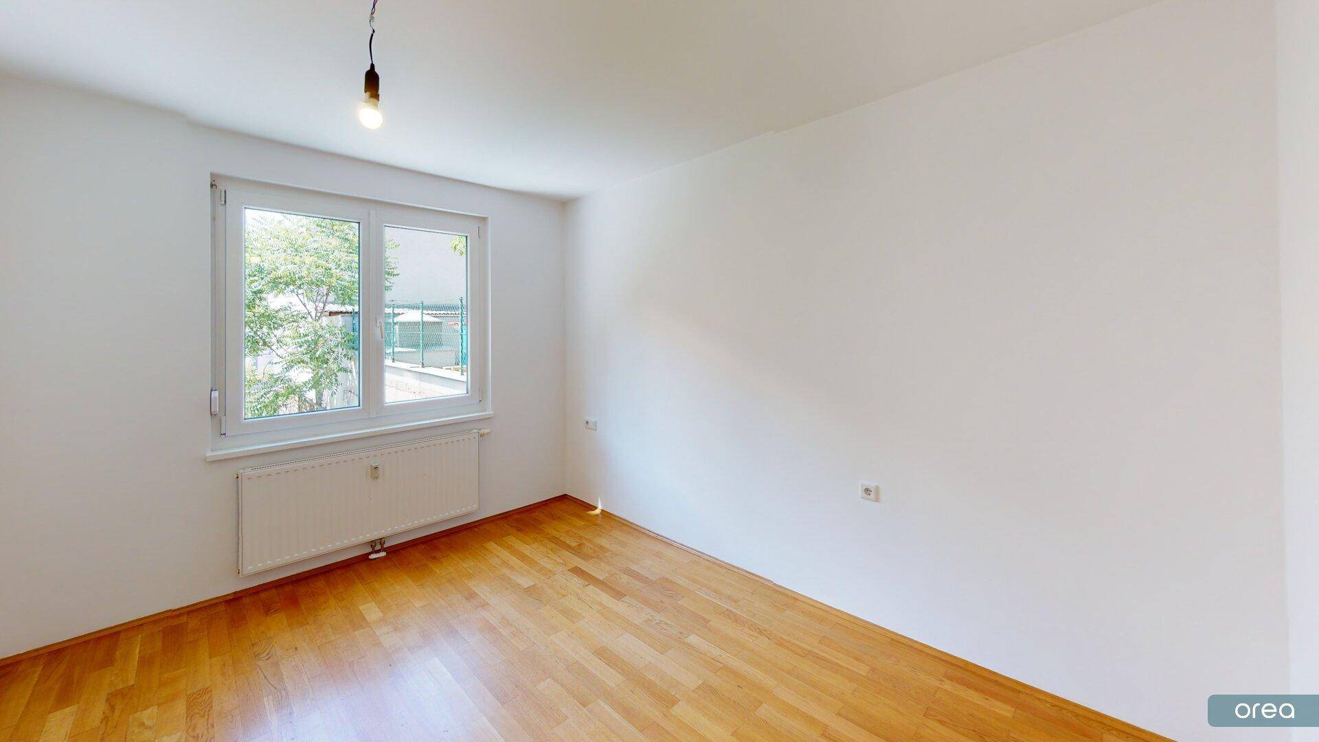 Helles Schlafzimmer mit Parkettboden, weißen Wänden und einem Fenster mit Blick ins Grüne.