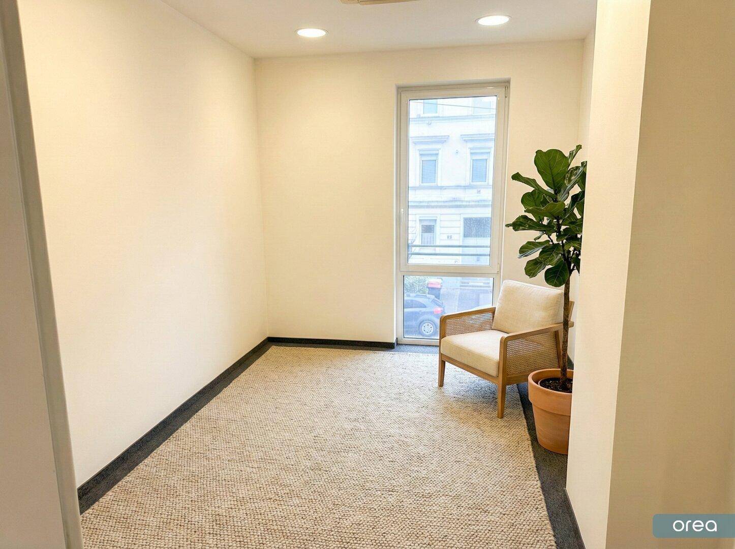 Kleiner, heller Raum mit Teppichboden, einem großen Fenster und einer Pflanze, geeignet als Einzelbüro oder Wartebereich.
