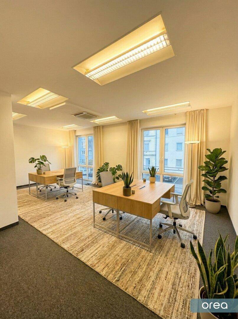 Großes, helles Büro mit mehreren Arbeitsplätzen, Teppichboden und großen Fenstern mit Blick nach draußen.