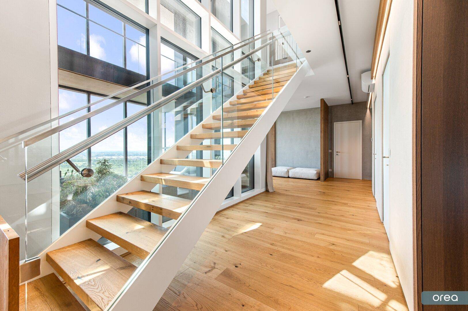 Heller Flur mit moderner Holztreppe, Glasgeländer und raumhohen Fenstern mit Panoramablick.