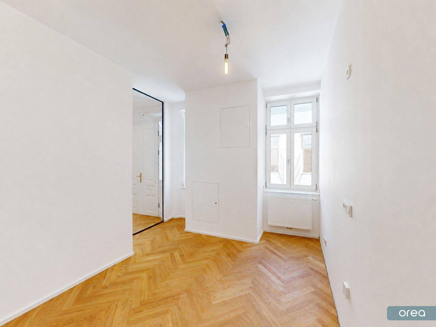 Helles Zimmer mit Fischgrätparkett, einem Fenster und einer modernen Spiegeltür.