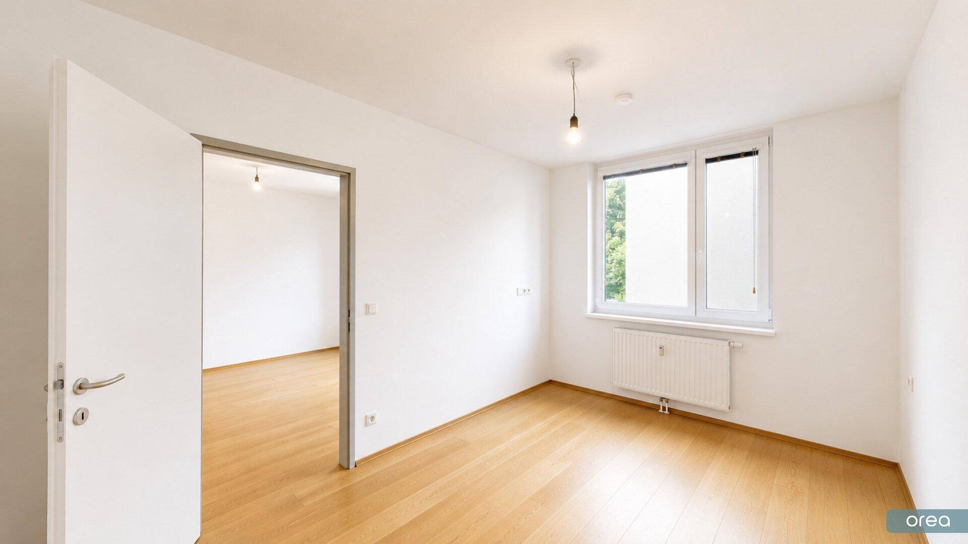 Leeres Schlafzimmer mit hellem Parkettboden und einem Fenster, das viel Tageslicht hereinlässt.
