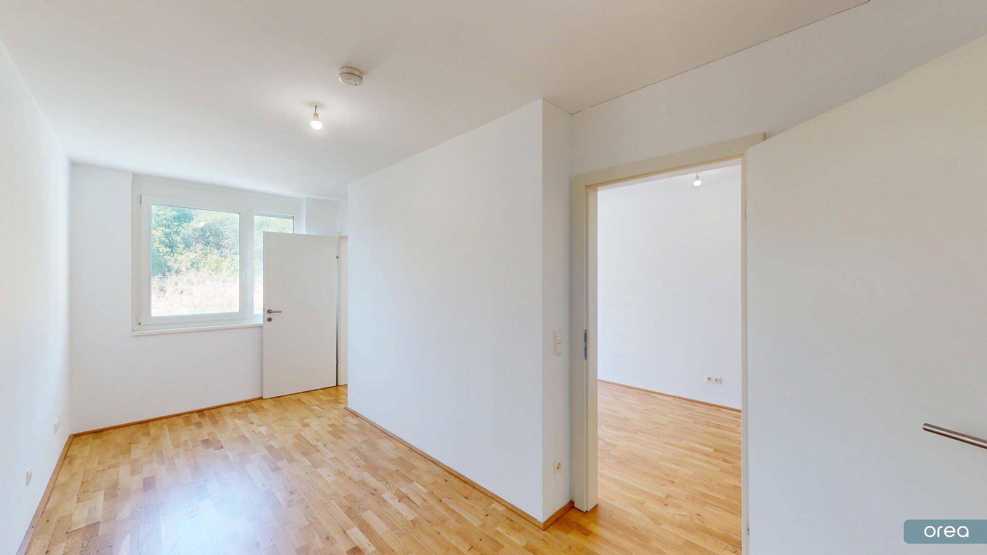 Leeres Schlafzimmer mit Holzboden und einem Fenster, das viel Tageslicht hereinlässt.