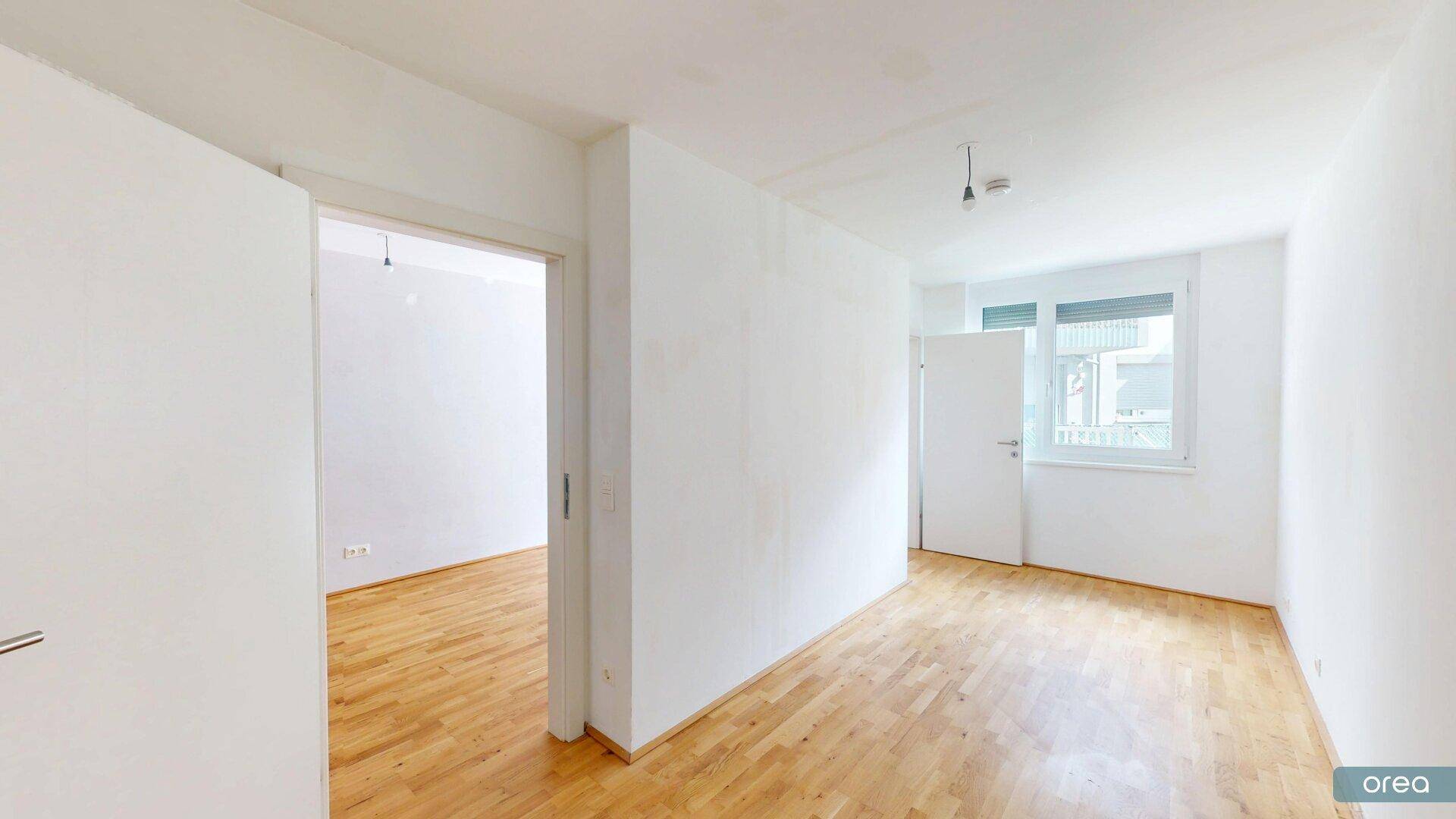 Heller Raum mit Holzboden und Fenster, der viel Tageslicht bietet und Zugang zu einem weiteren Zimmer.