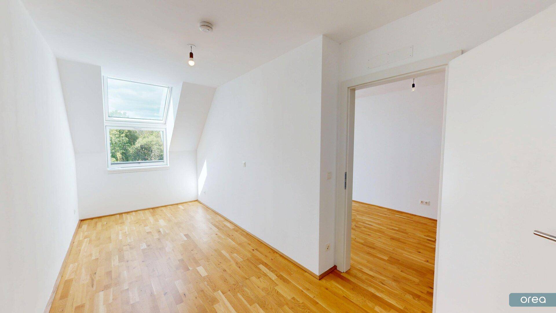 Helles Zimmer mit Holzboden, Dachschräge und großem Dachfenster mit Blick auf Bäume.