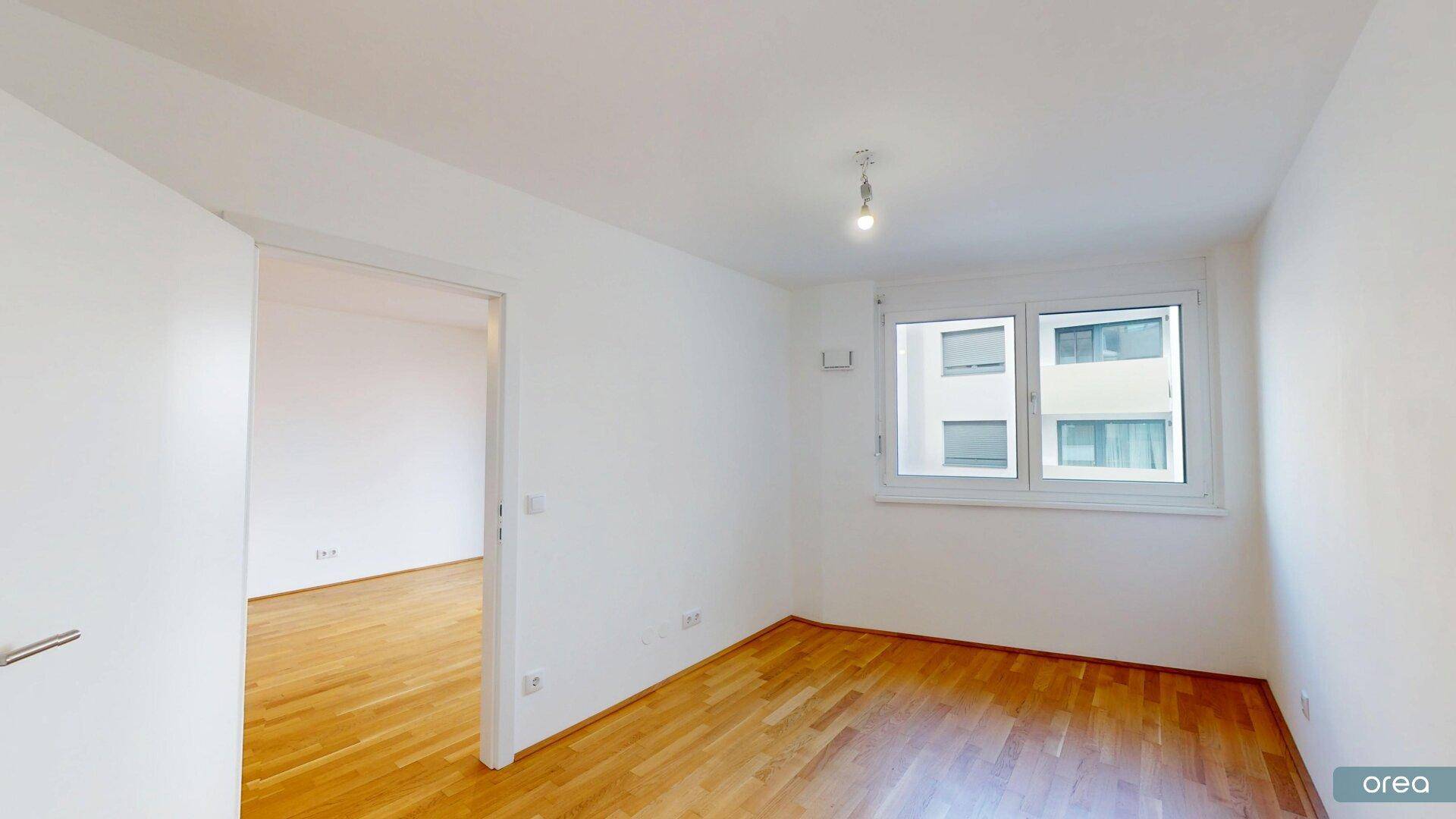 Helles Schlafzimmer mit Holzboden und einem Fenster, das den Blick auf die umliegenden Gebäude freigibt.