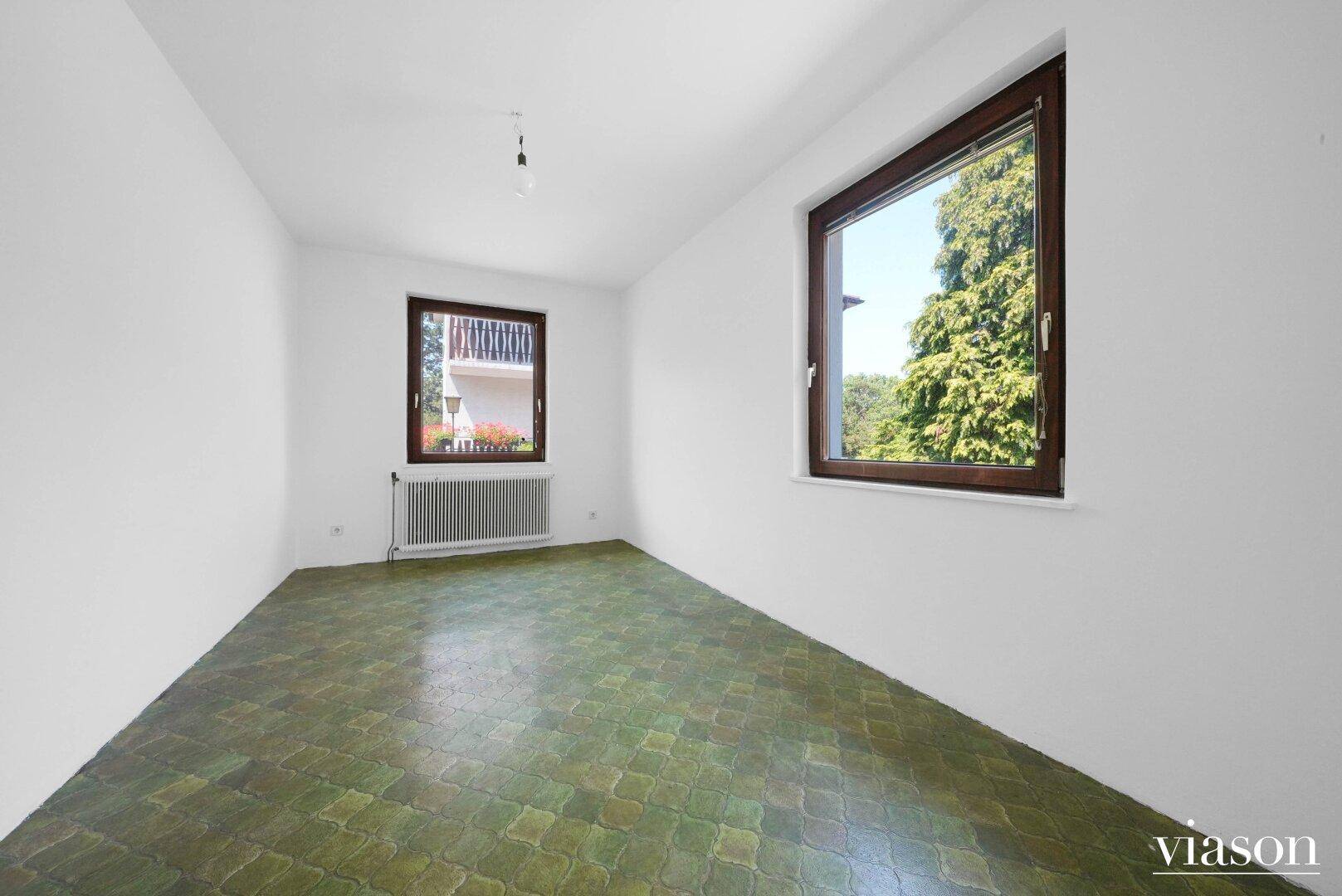 Schlafzimmer mit grünem Fliesenboden und zwei Fenstern mit Blick ins Grüne.