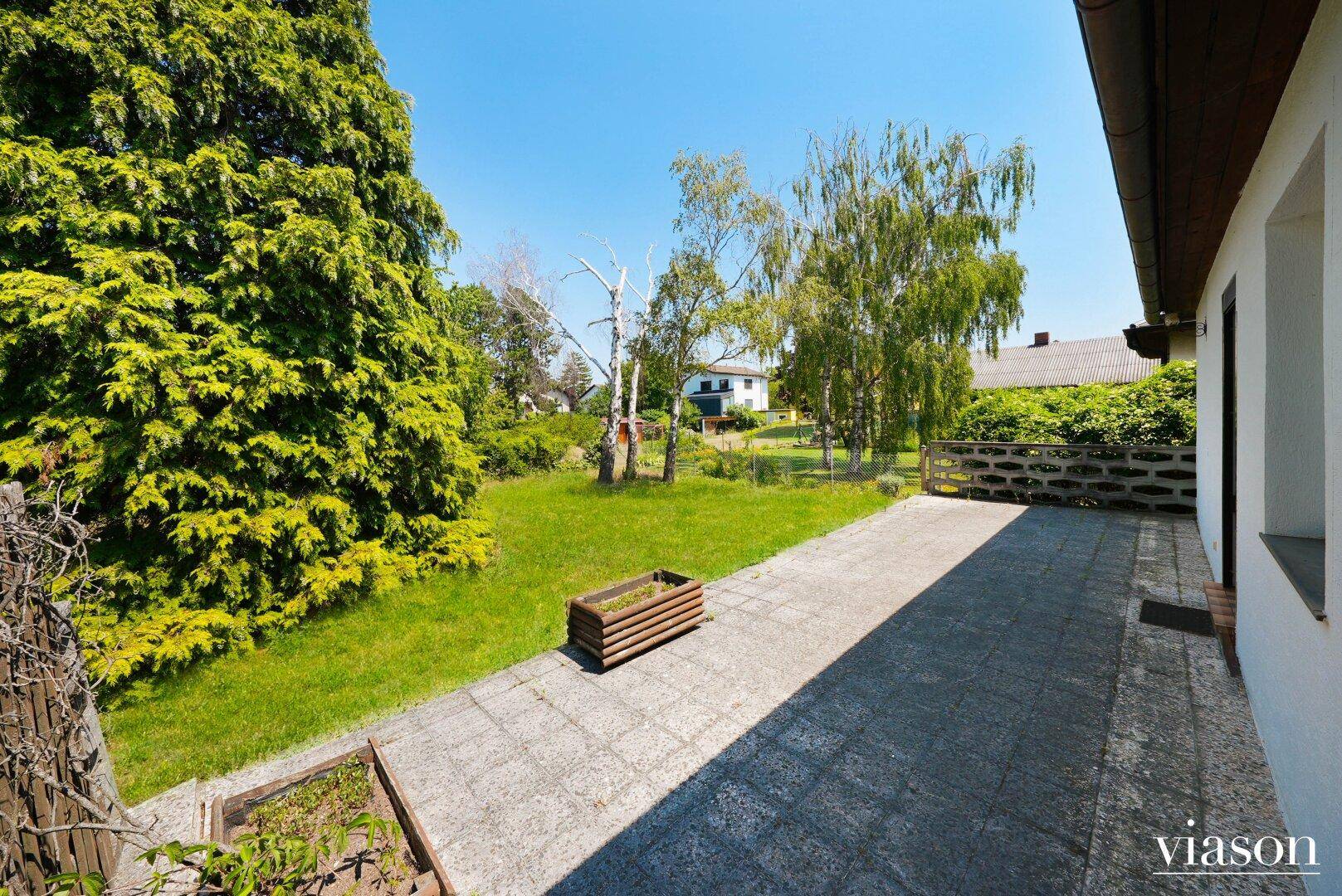 Große gepflasterte Terrasse mit angrenzendem Rasenbereich und viel Grün im Garten.
