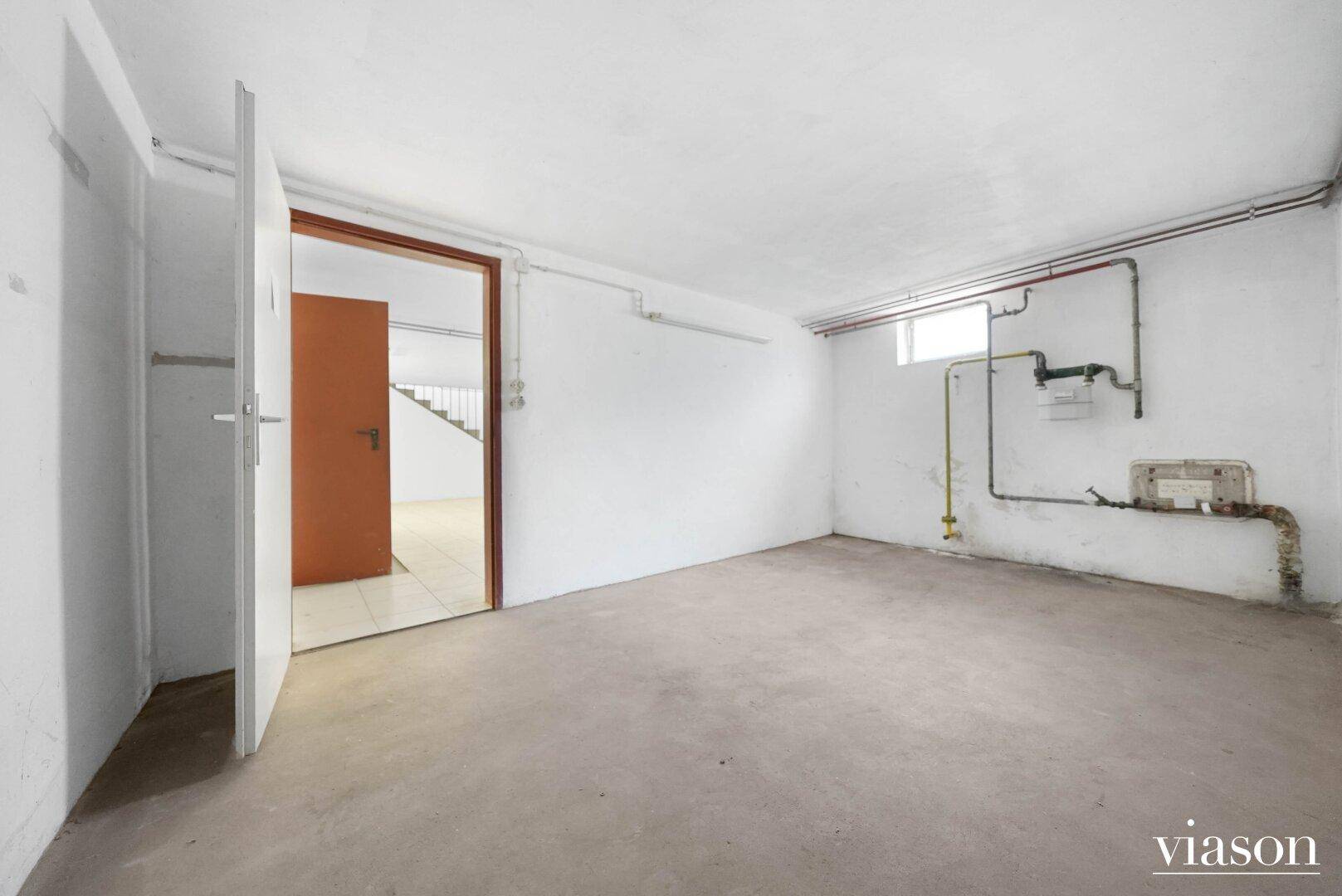 Kellerraum mit sichtbaren Rohren, Fenster und Zugang zu weiteren Räumen.