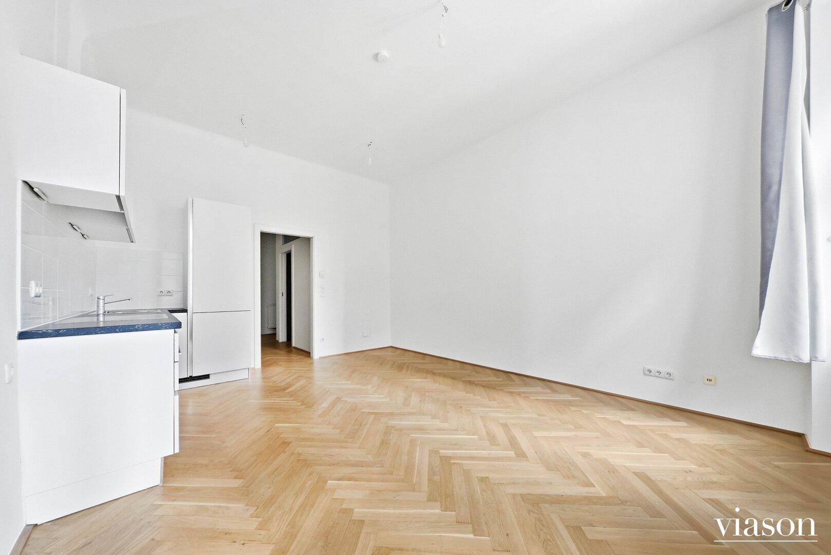 Leere Studio-Wohnung mit Fischgrätparkett, weißen Wänden und einer integrierten Küchenzeile.