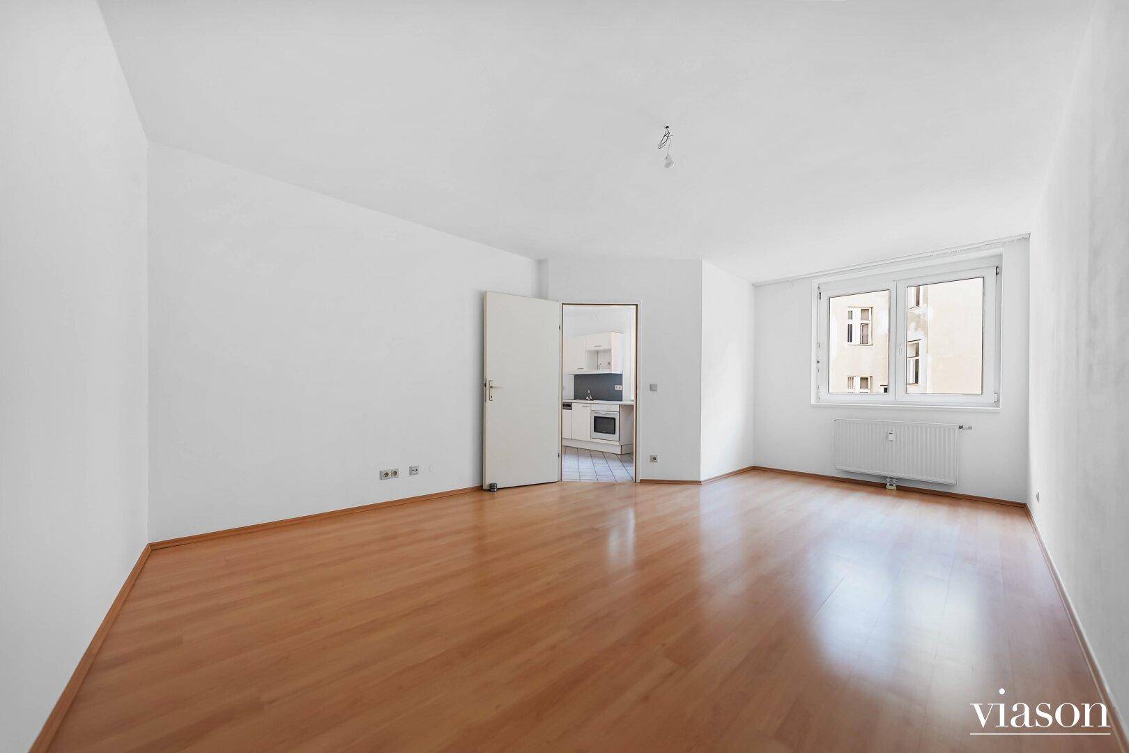 Helles, leeres Zimmer mit Parkettboden, weißen Wänden und Blick nach draußen.