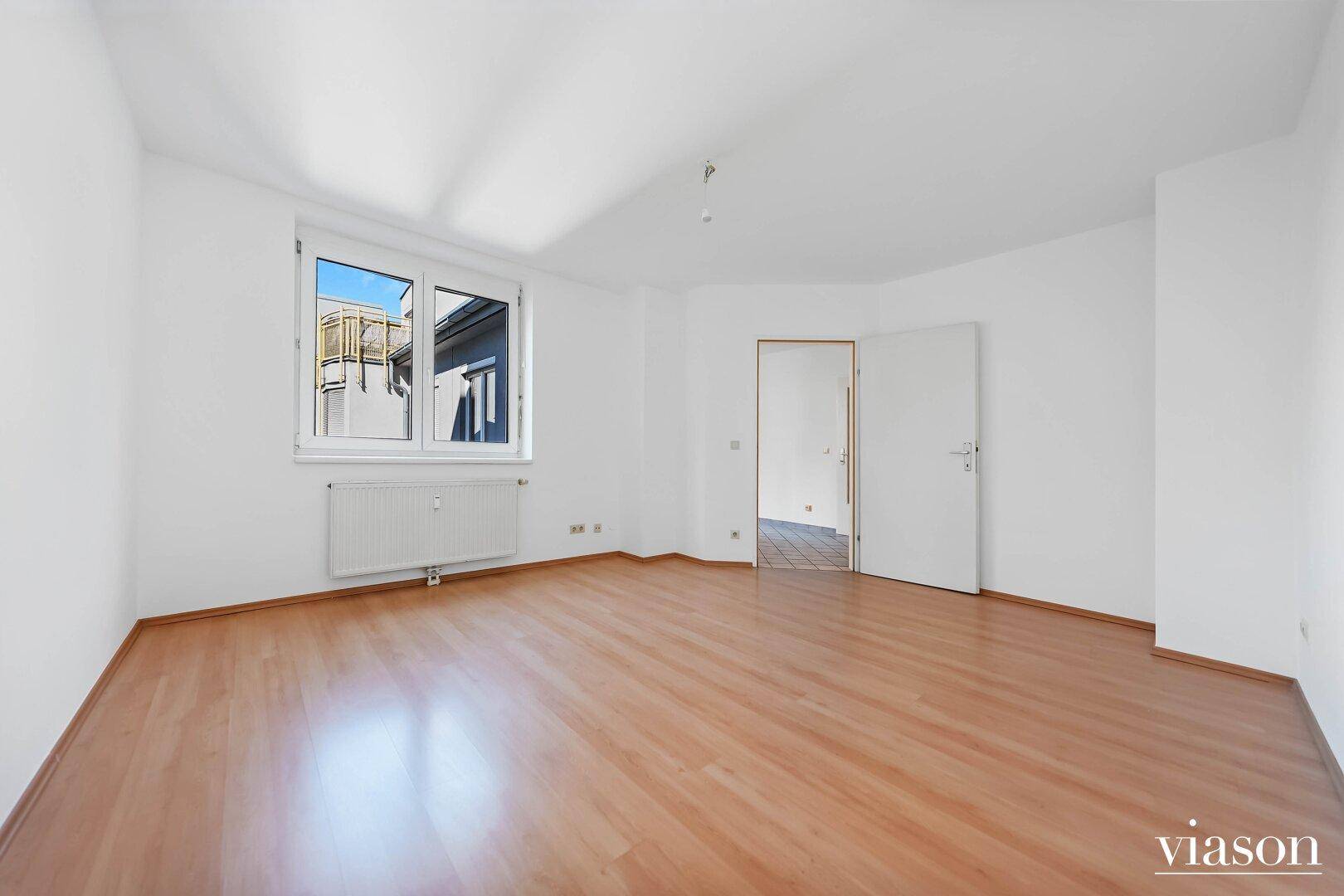 Helles, leeres Zimmer mit Parkettboden, weißen Wänden und einem Fenster mit Ausblick.