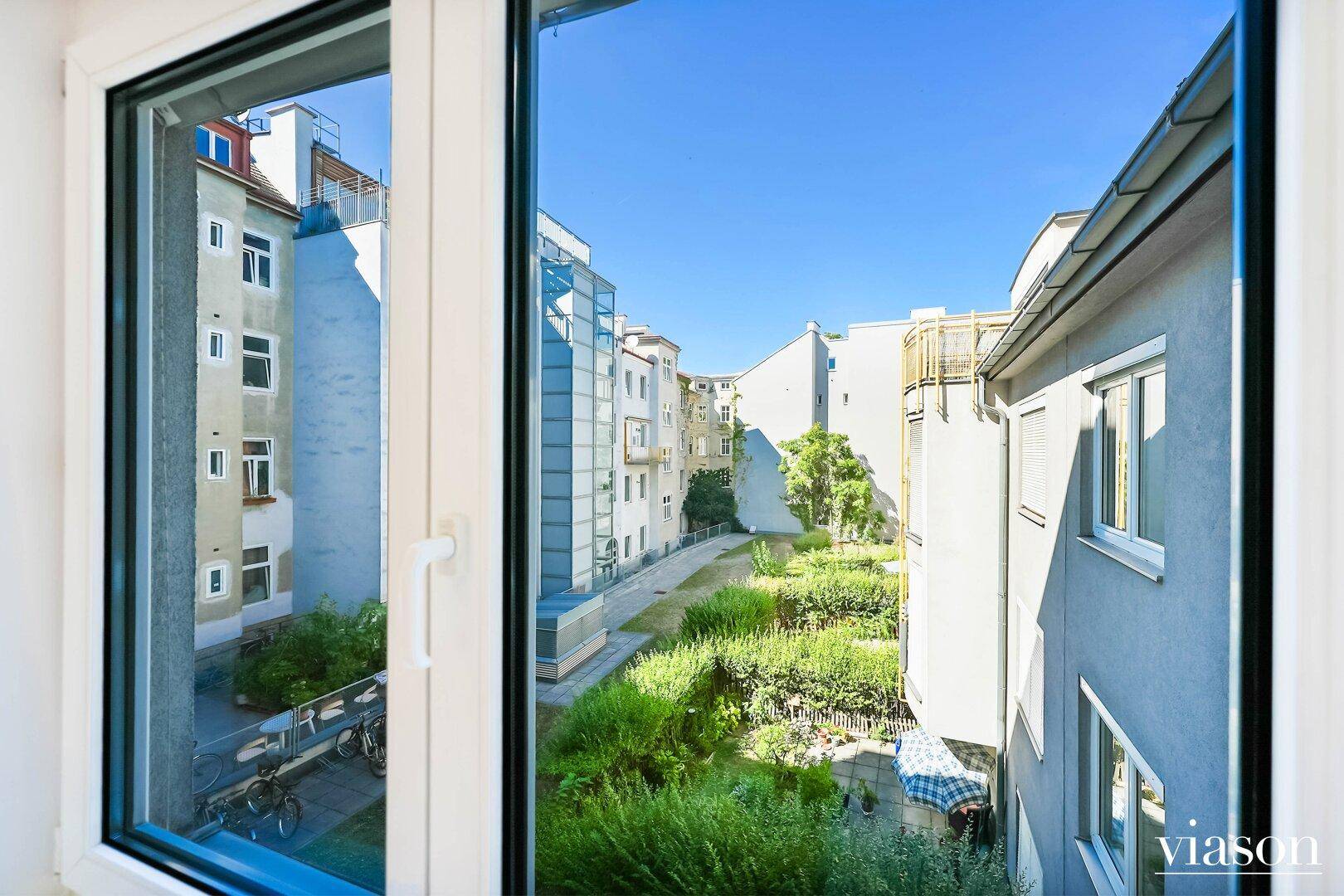 Blick aus dem Fenster auf einen grünen Innenhof mit umliegenden Gebäuden bei Sonnenschein.