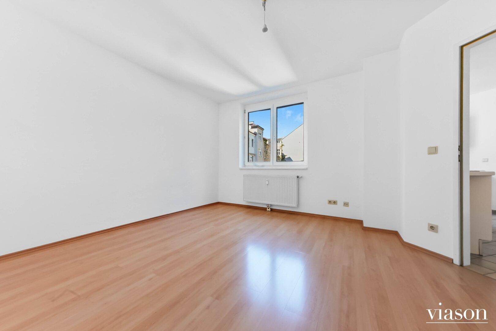 Helles, leeres Zimmer mit Parkettboden, weißen Wänden und einem Fenster mit Ausblick.