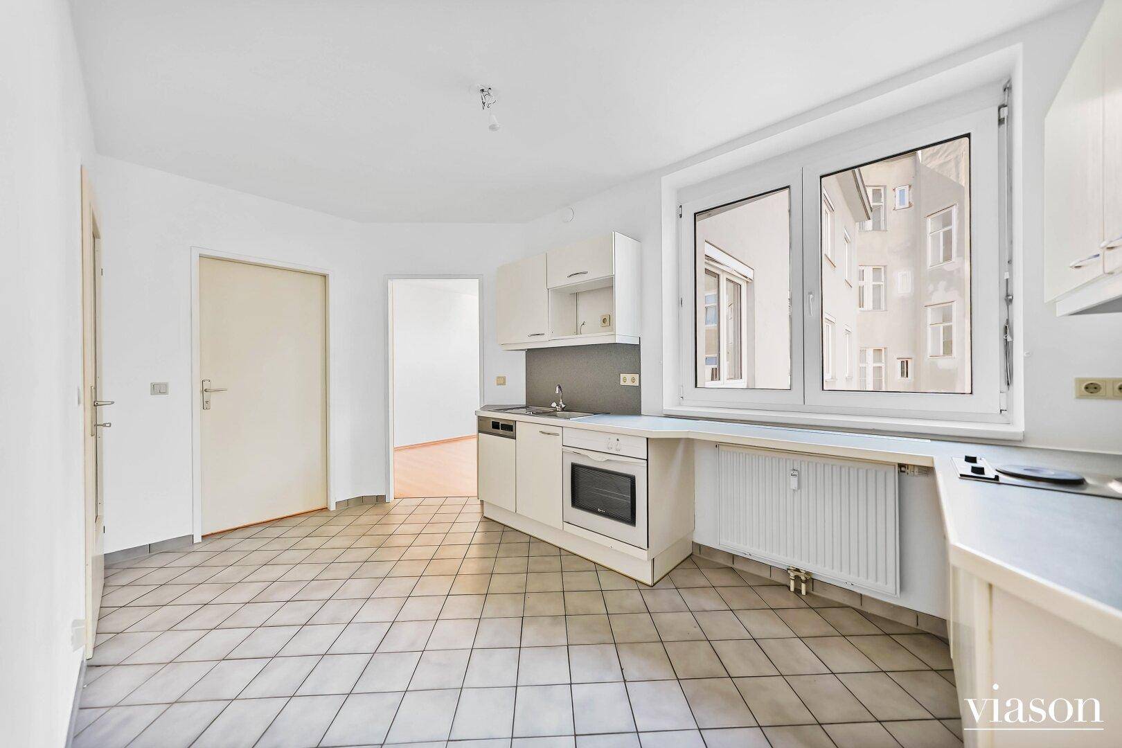 Funktionale Küche mit weißen Schränken, Fliesenboden und einem Fenster mit Heizkörper darunter.