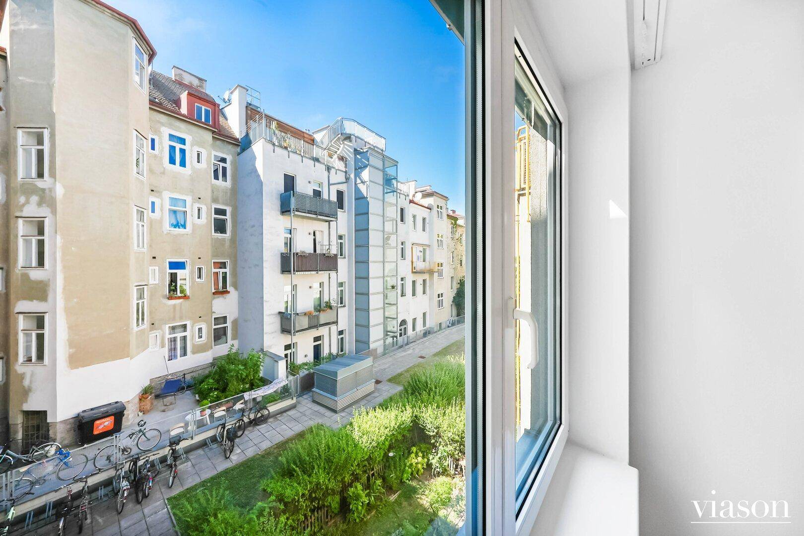 Weitblick aus dem Fenster auf den Innenhof mit viel Grün, Fahrrädern und einem externen Glasaufzug.