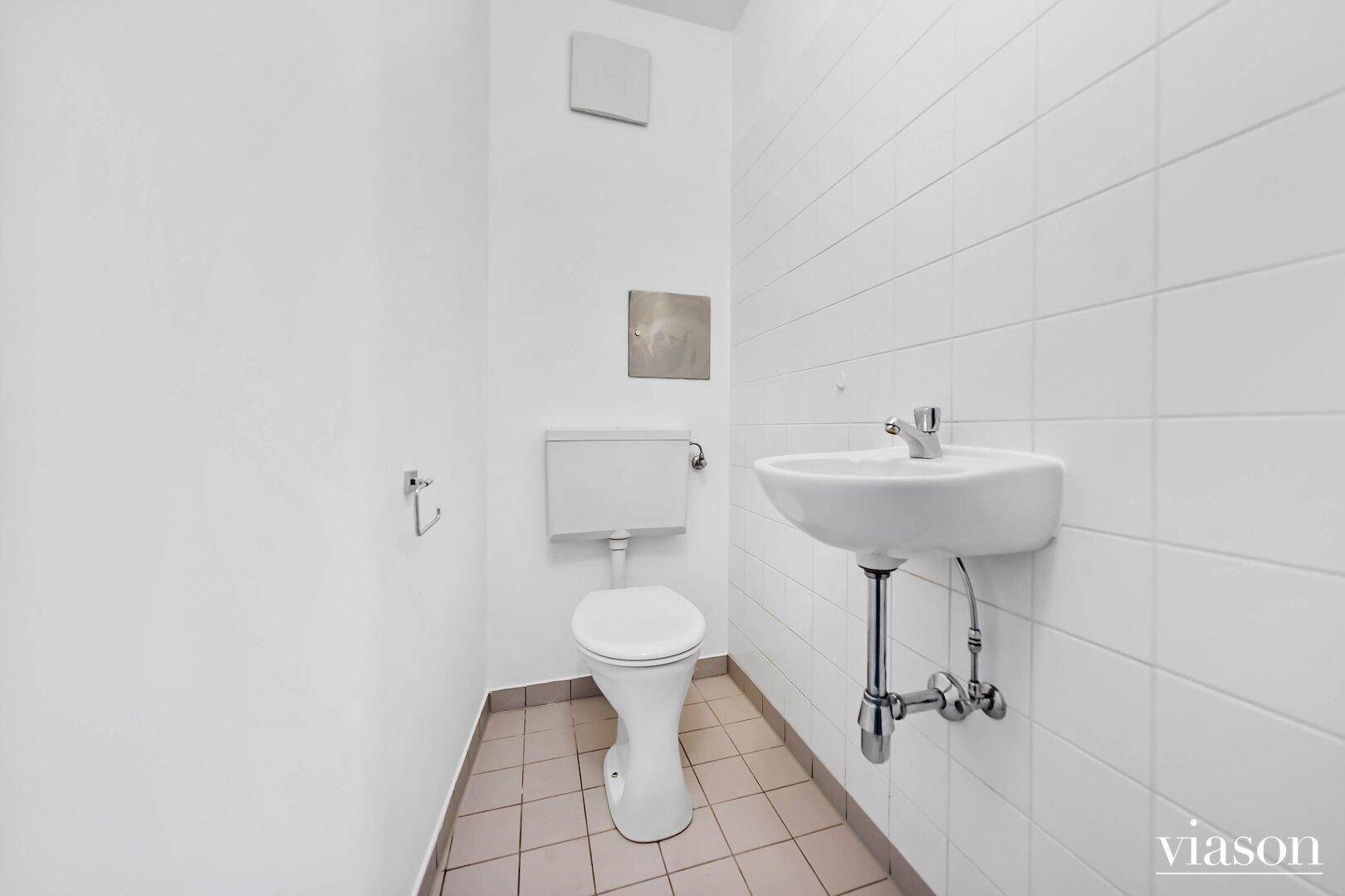 Separates WC mit Toilette, kleinem Handwaschbecken und weißen Wand- und Bodenfliesen.