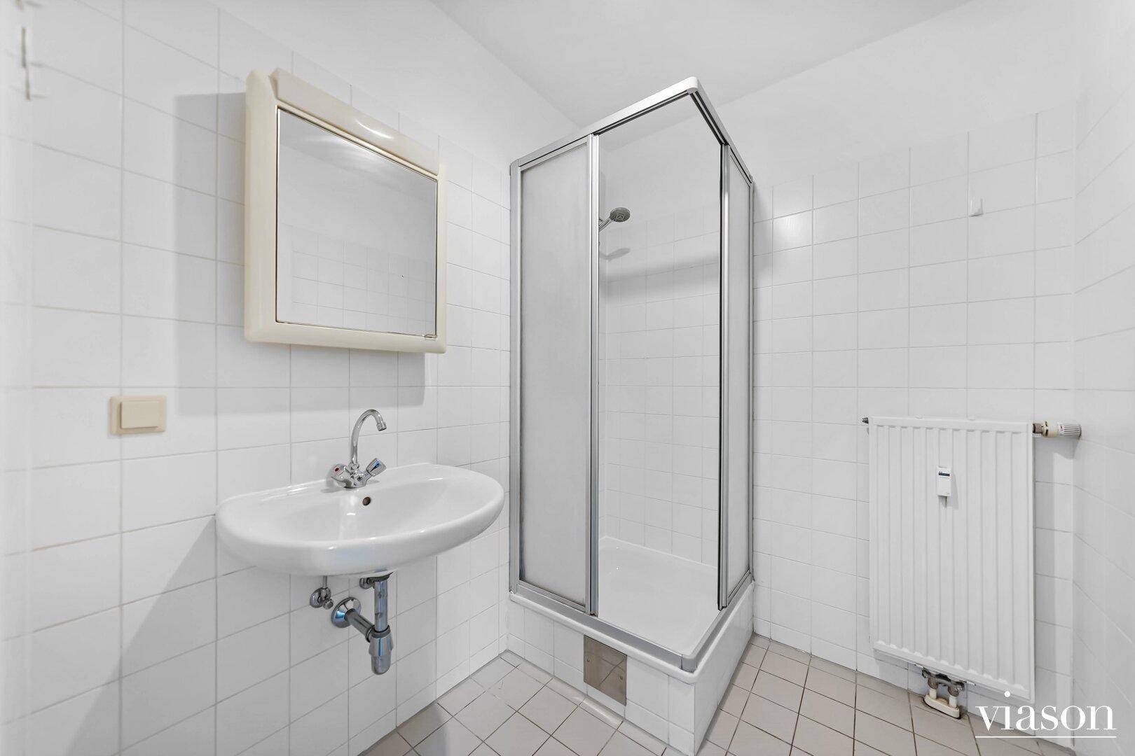 Helles Badezimmer mit weißen Fliesen, Waschbecken, Spiegelschrank und einer Duschkabine.