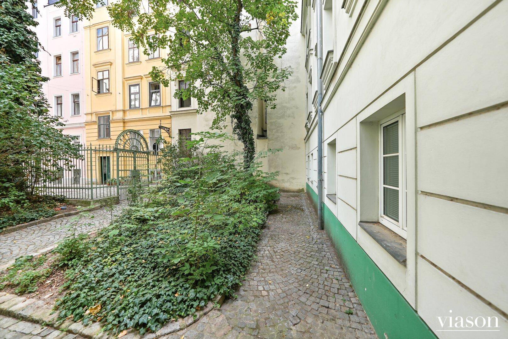Schmaler Weg im begrünten Innenhof, umgeben von Altbaufassaden und Pflanzen.
