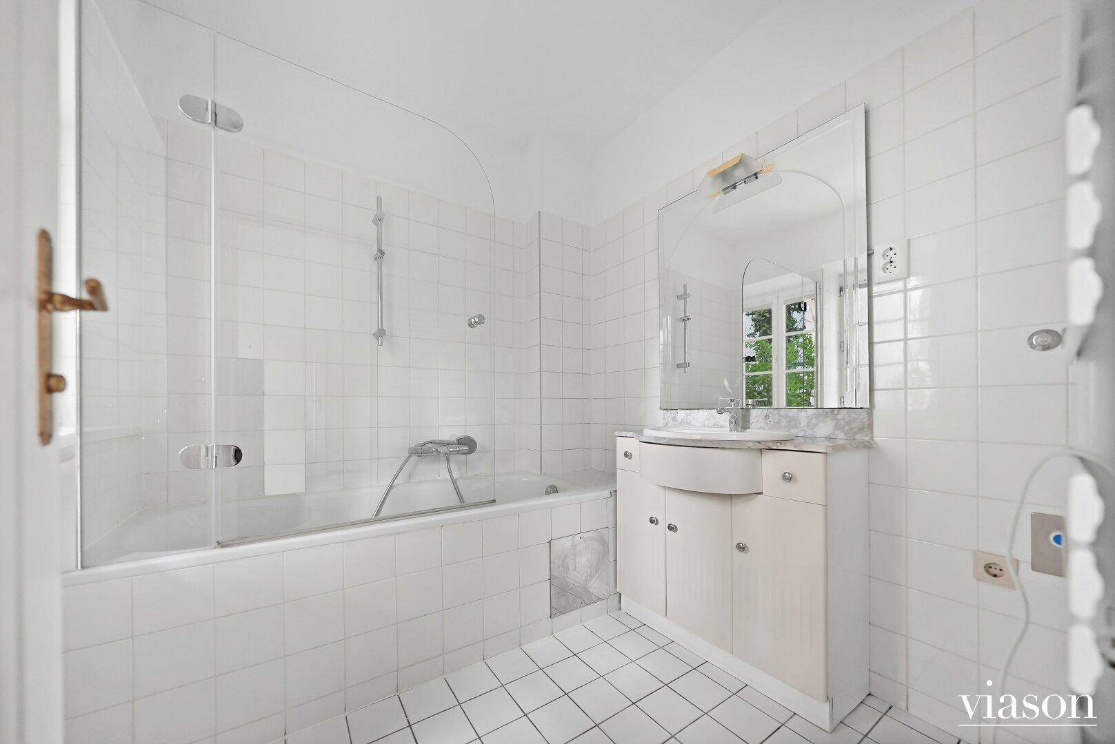 Helles Badezimmer mit weißen Fliesen, Badewanne mit Duschwand und Waschtisch mit Marmorplatte.